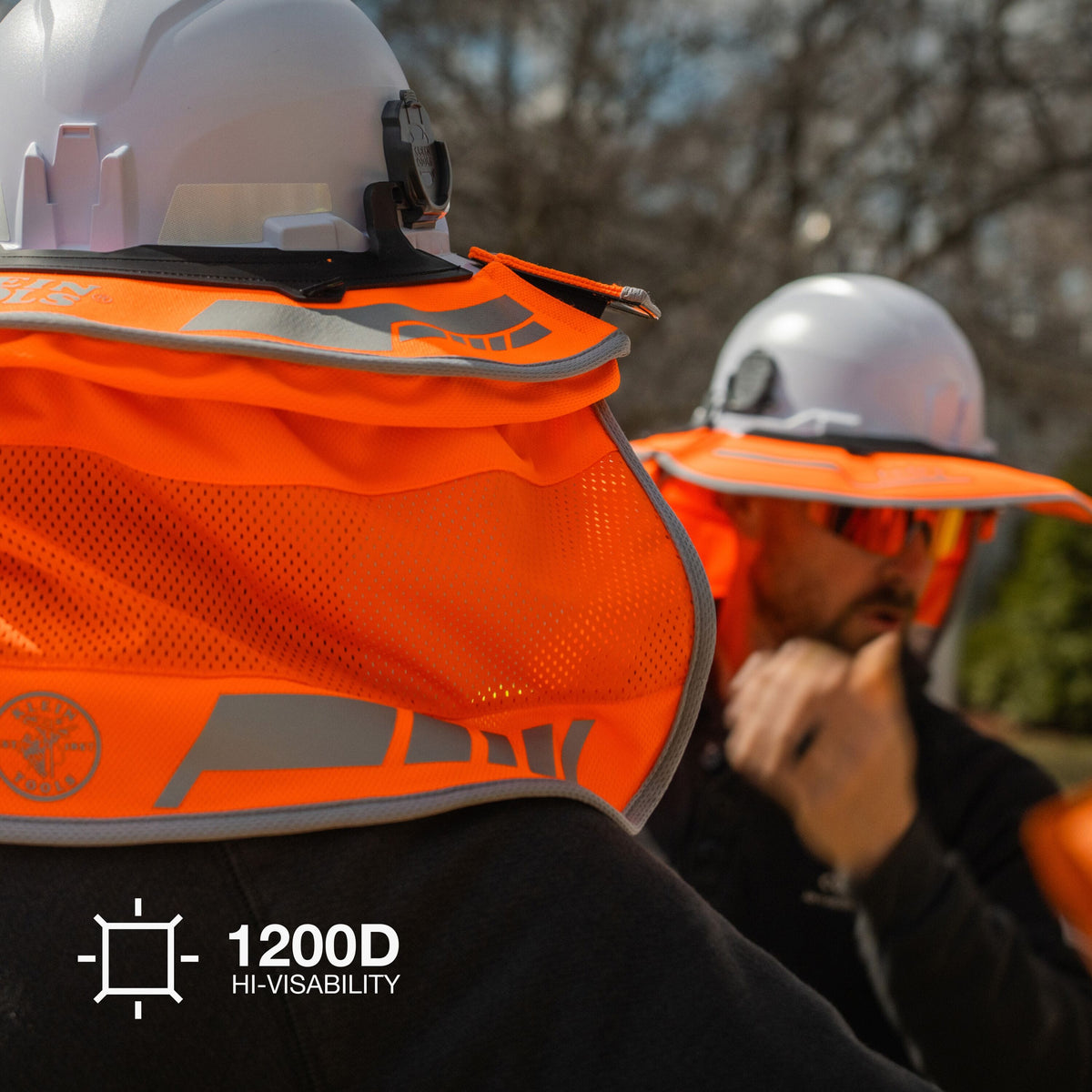 Close-up of a high-visibility orange mesh hard hat cover worn by a construction worker outdoors, with another worker in the background and a '1200D HI-VISIBILITY' badge in the lower left corner.