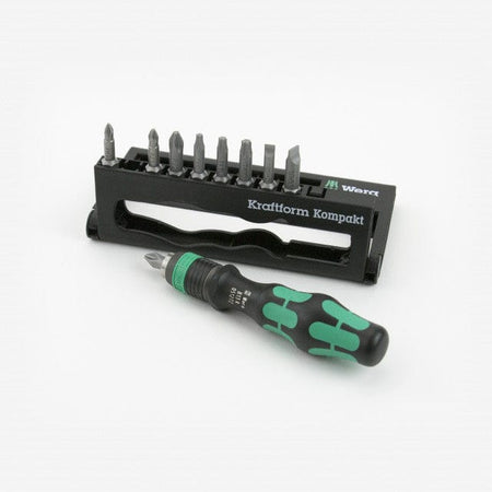 Wera - 5056653001 KK 10 Compact Bitholder and Bit Set, 10-Piece: This image depicts a set of tools laid out on a light-colored surface. A black plastic holder, standing upright, displays a row of silver-colored screwdriver bits along its top edge, while a screwdriver handle with a black and green grip lies horizontally in the foreground. The brand name "Wera" is visible on both the holder and the handle.