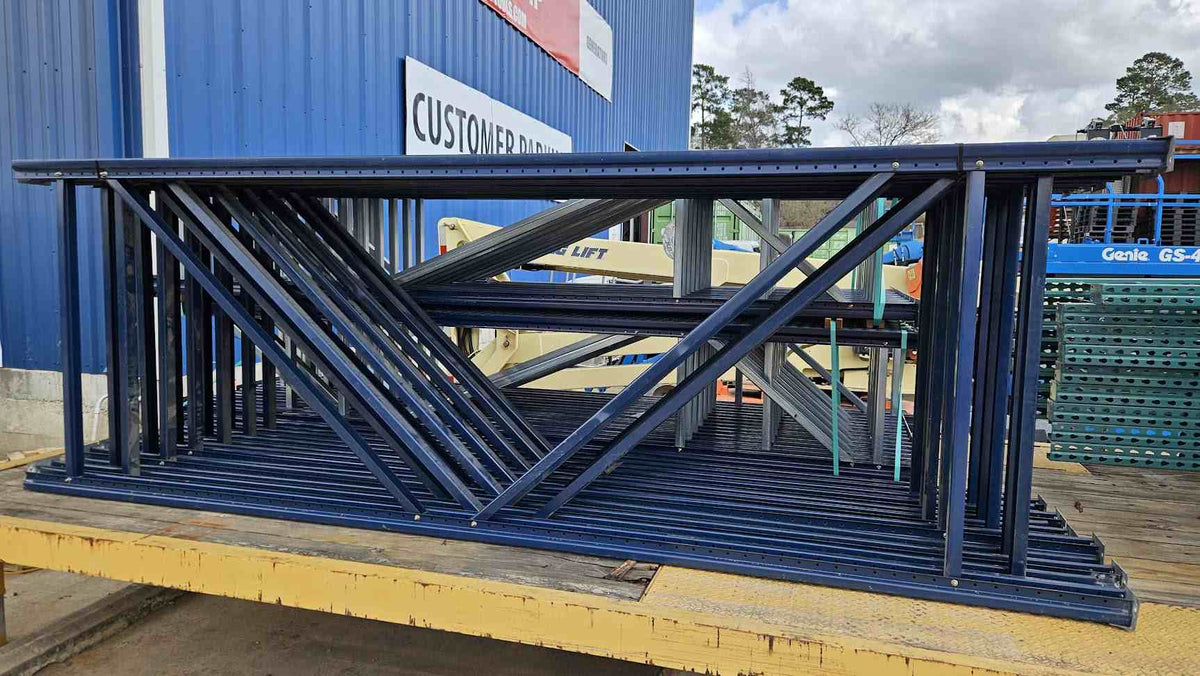 A horizontal stack of used like-new 12-foot industrial blue pallet rack uprights, featuring heavy-duty steel frames with a 48-inch depth, organized on a flatbed trailer for high-capacity warehouse storage system transport.