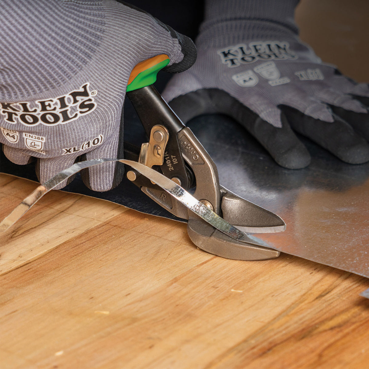 Klein Tools tin shears cutting a sheet of metal on a wooden workbench, with a person wearing gray protective gloves holding the tool and glove.