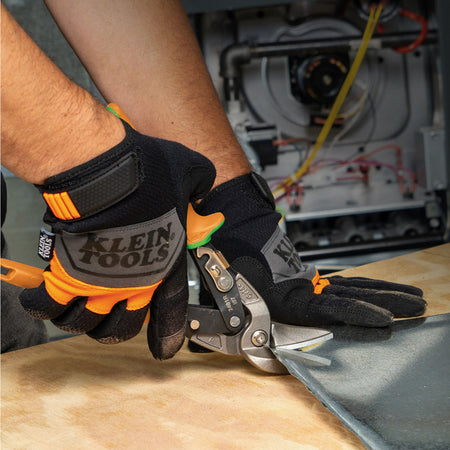 Close-up of a person wearing black and orange Klein Tools work gloves while using metal shears on a sheet of metal over a wood surface, with a blurred workshop background.