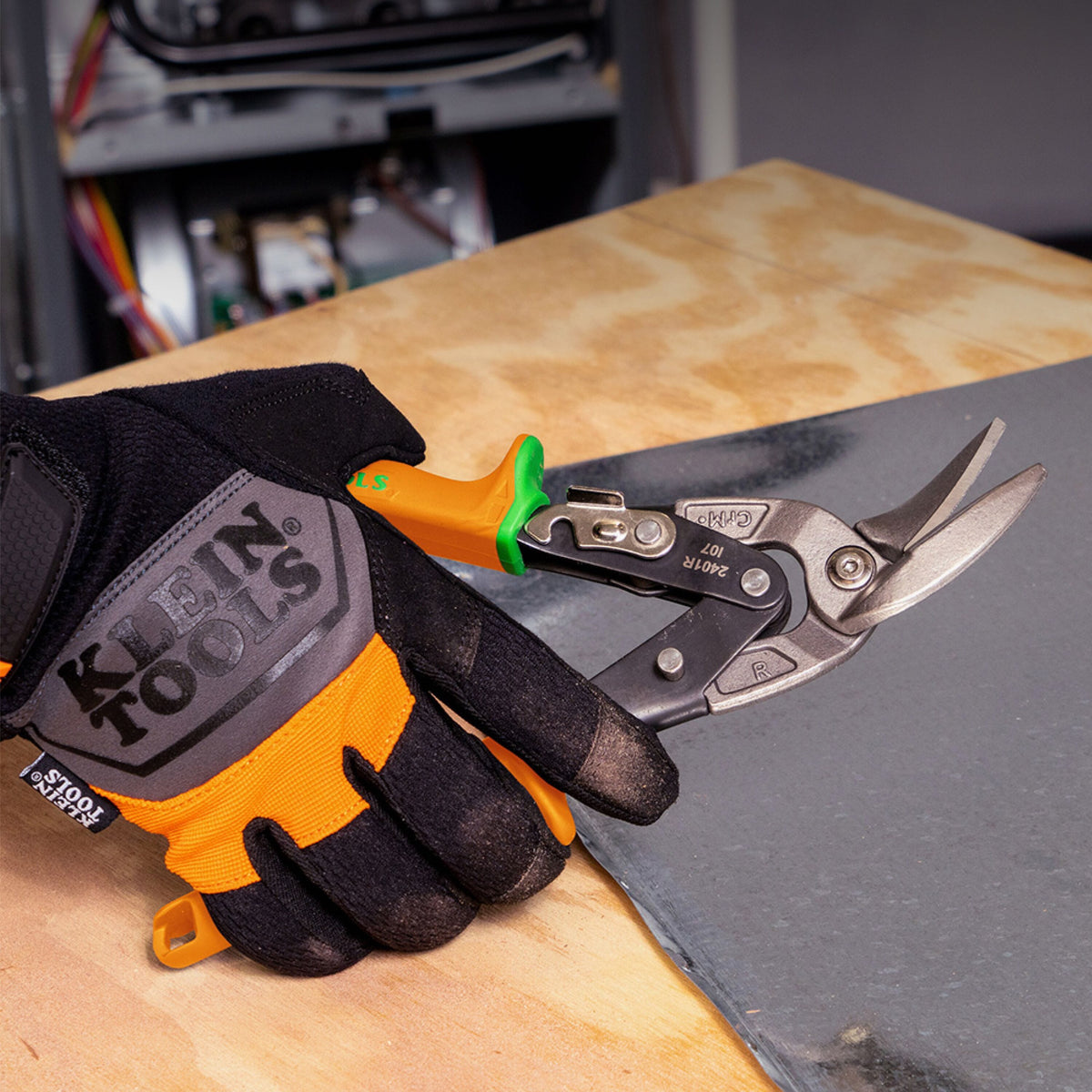 A hand wearing a black and orange Klein Tools work glove holds a pair of metal tin shears with orange and green handles over a wooden workbench next to a dark gray sheet.