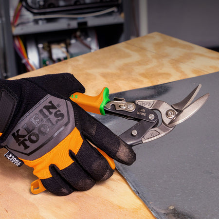 A hand wearing a black and orange Klein Tools work glove holds a pair of metal tin shears with orange and green handles over a wooden workbench next to a dark gray sheet.