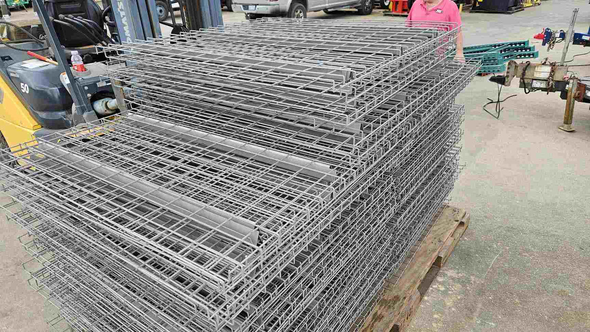 A large stack of used like-new 48-inch by 46-inch pallet rack wire decking panels made of galvanized steel mesh, organized on a wooden pallet next to a yellow forklift for industrial warehouse storage and fire safety compliance.