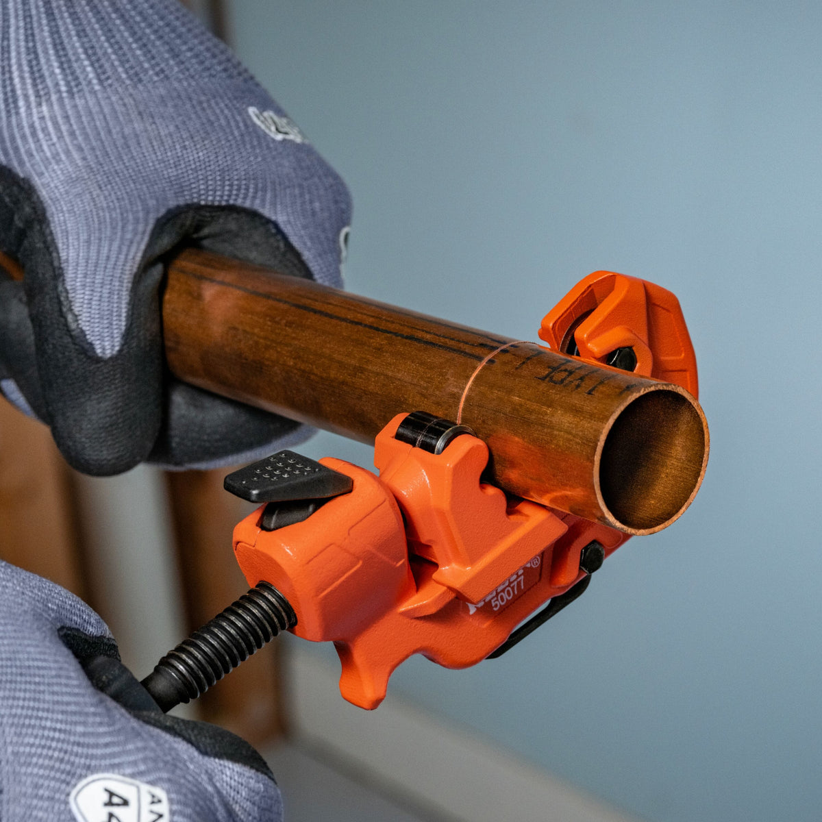 An orange industrial clamp holding a copper pipe section, with a person wearing gray knit work gloves gripping both ends against a pale blue background.