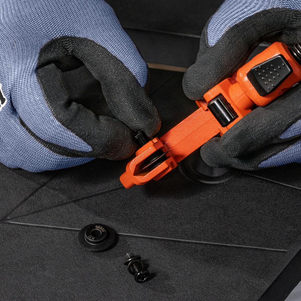 Close-up of gloved hands using an orange and black plastic tool to remove a small black fastener from a dark textured surface.