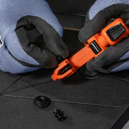 Close-up of gloved hands using an orange and black plastic tool to remove a small black fastener from a dark textured surface.