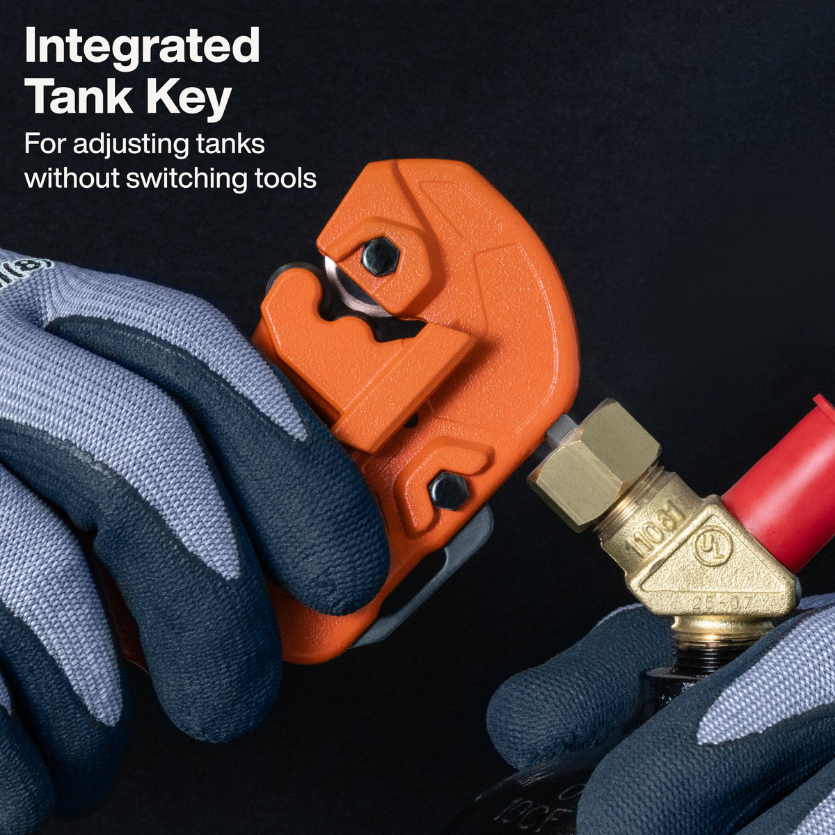 Close-up of a person wearing gray and blue work gloves using an orange integrated tank key tool to adjust a brass pipe fitting with a red hose. White text in the upper left reads 'Integrated Tank Key' and 'For adjusting tanks without switching tools.