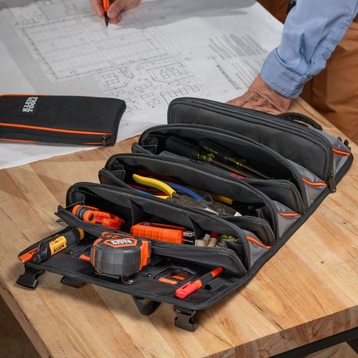 A black and gray multi-compartment tool organizer pouch with orange accents, shown open on a wooden workbench with various hand tools inside. A person's hand and arm are visible in the background near blueprints, and a matching closed pouch is visible on the left.