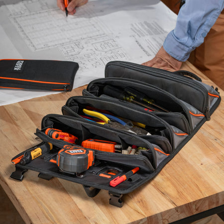 A black and gray multi-compartment tool organizer pouch with orange accents, shown open on a wooden workbench with various hand tools inside. A person's hand and arm are visible in the background near blueprints, and a matching closed pouch is visible on the left.