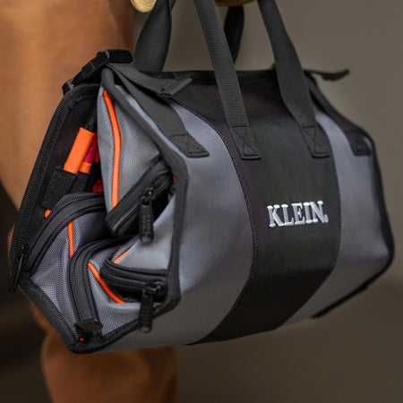 A close-up of a black and gray KLEIN duffel bag being held by a person, showing its multiple zippered compartments and bright orange interior lining.