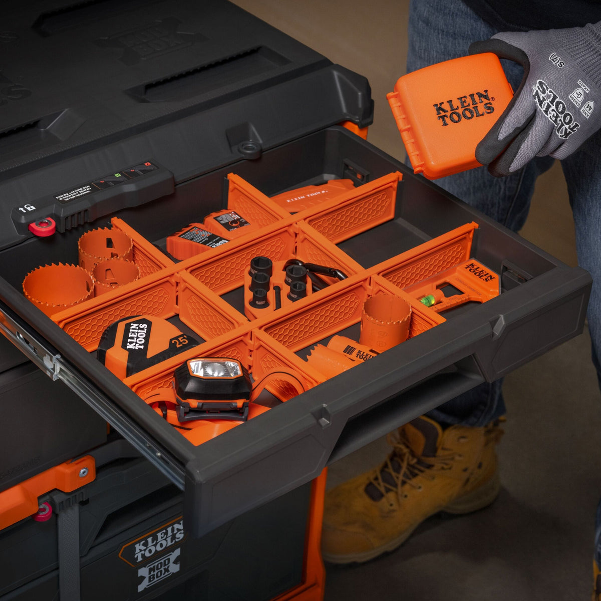 A person wearing gloves holds an orange Klein Tools storage organizer tray next to an open black and orange Klein Tools tool box filled with organized power tools and accessories.