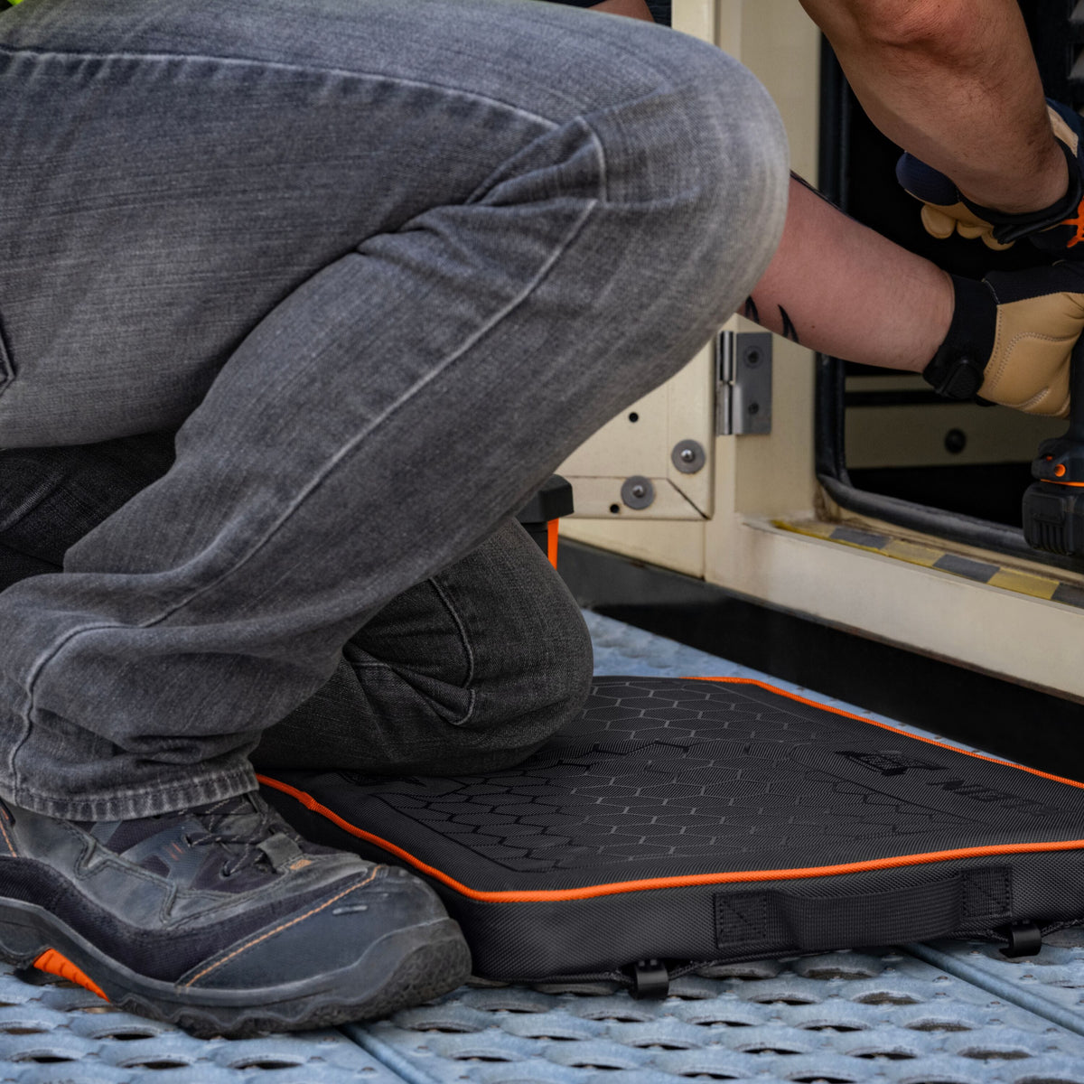 A black rubber vehicle floor mat with orange trim is shown on a metal grate near an open vehicle door, with a person kneeling beside it and another person's arm reaching in. The mat has a textured surface and small black feet at the bottom.