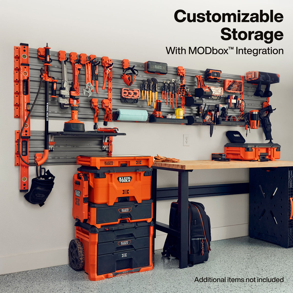 A workshop wall-mounted tool organizer with orange and black power tools arranged on a gray slatwall, alongside orange and black storage bins and a workbench. Text at the top reads 'Customizable Storage With MODbox Integration' and text at the bottom right says 'Additional Items not included.