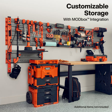 A workshop wall-mounted tool organizer with orange and black power tools arranged on a gray slatwall, alongside orange and black storage bins and a workbench. Text at the top reads 'Customizable Storage With MODbox Integration' and text at the bottom right says 'Additional Items not included.