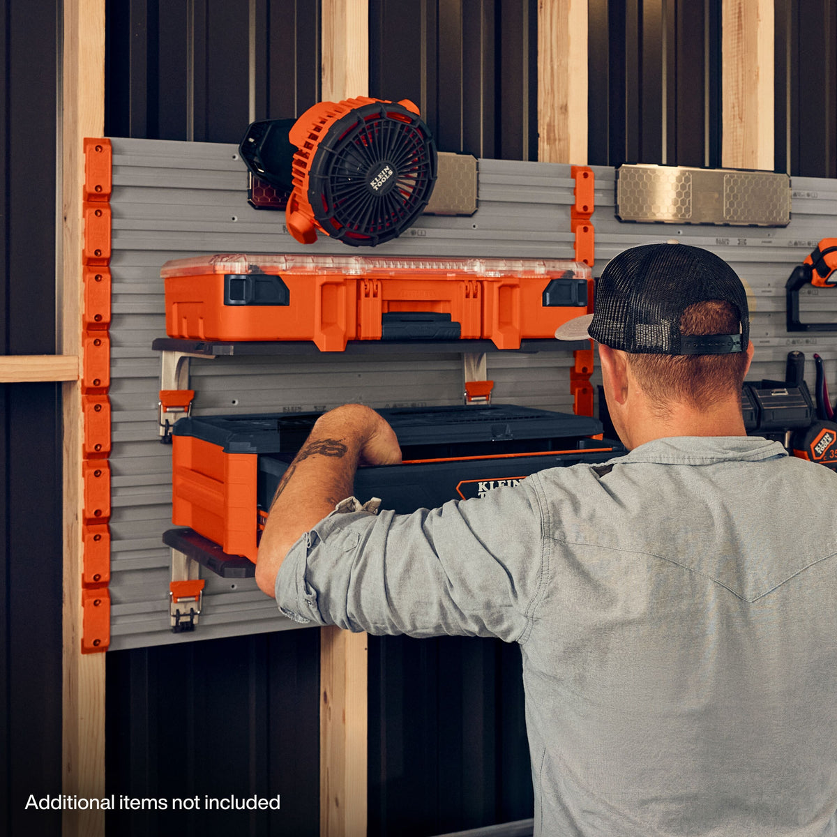 A man in a gray shirt and black cap works on a gray wall-mounted tool organizer panel with orange brackets, shelves, and tools, including a black and orange circular fan-like tool and a black storage case. The scene is set in a workshop with wood framing visible.