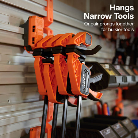 Bright orange and black GCL clamp tools hanging on a gray garage door wall, with white text in the upper right reading 'Hangs Narrow Tools Or pair prongs together for bulkier tools.'.