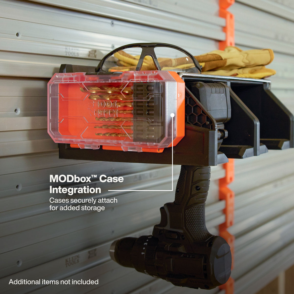 A black wall-mounted tool organizer labeled MODbox Case Integration, shown attached to a gray garage door. The organizer holds a translucent orange-and-black storage case with drill bits and a pair of tan gloves on a black shelf, with a drill hanging below.