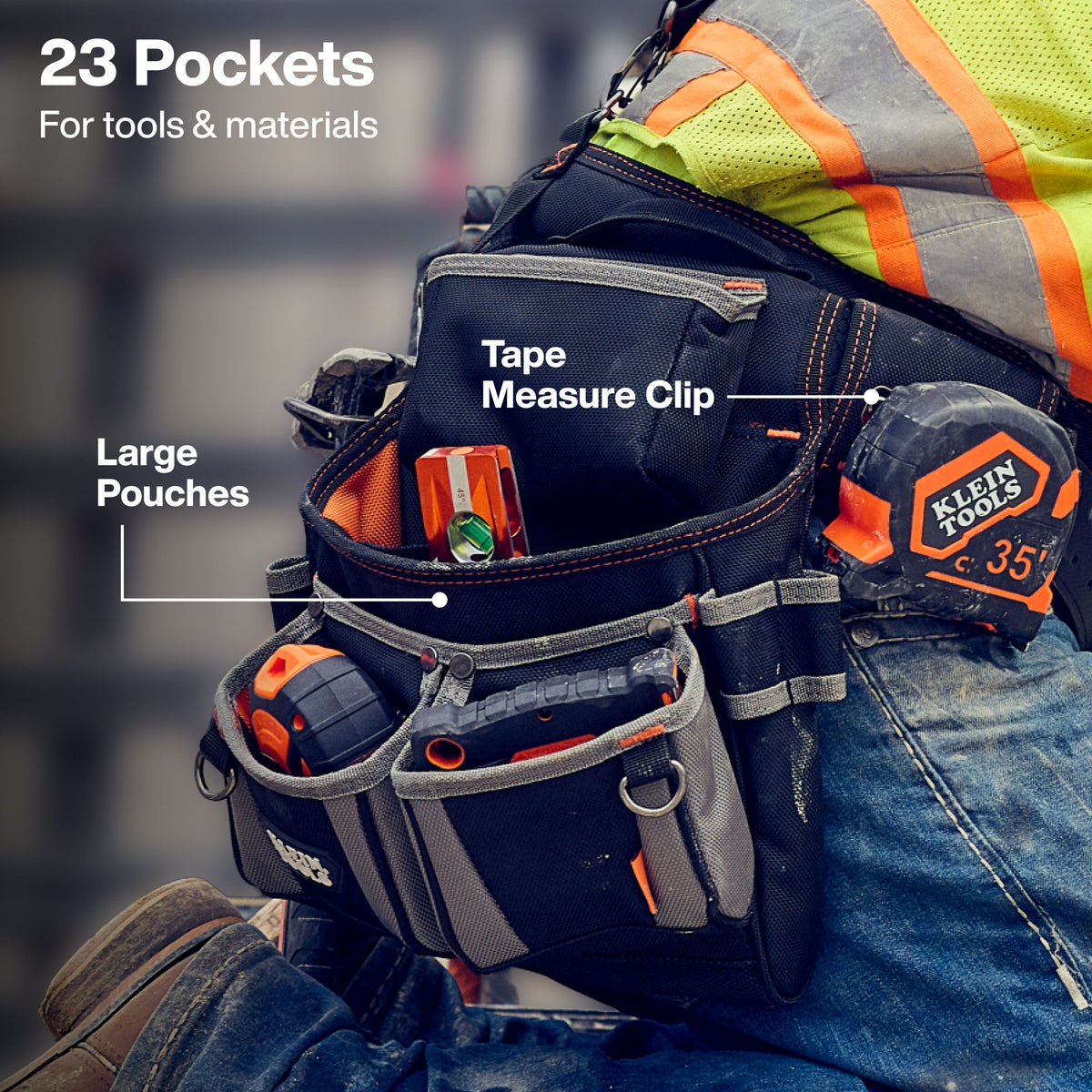 The shot focuses on the worker's left side and thigh from a low angle. The worker is clad in blue denim jeans, a high-visibility safety vest with bright yellow fabric and orange/silver reflective bands, and heavy-duty work footwear. Suspended from their waist is a black heavy-duty ballistic polyester tool pouch assembly filled with measuring and layout equipment.