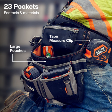 The shot focuses on the worker's left side and thigh from a low angle. The worker is clad in blue denim jeans, a high-visibility safety vest with bright yellow fabric and orange/silver reflective bands, and heavy-duty work footwear. Suspended from their waist is a black heavy-duty ballistic polyester tool pouch assembly filled with measuring and layout equipment.