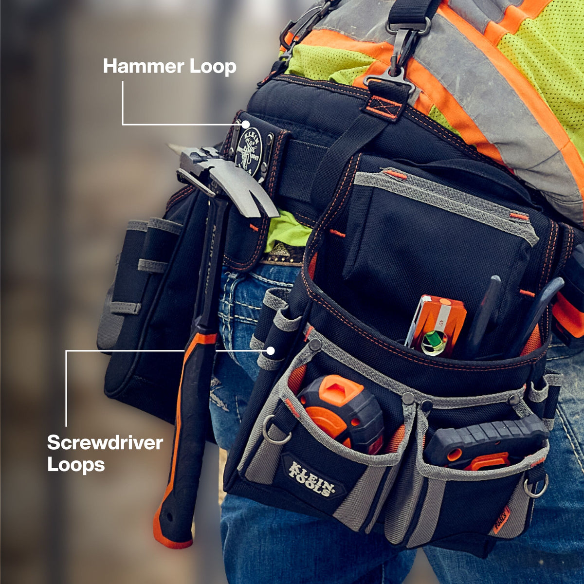 The worker is turned slightly away from the camera, wearing blue denim jeans, a high-visibility lime-yellow safety shirt, and an orange/silver reflective vest. Strapped securely at their waist is a black and gray ballistic-weave tool pouch system supported from above by a heavy-duty suspender harness with metal clip hardware. The rig is stocked with hand tools, layout equipment, and a claw hammer.