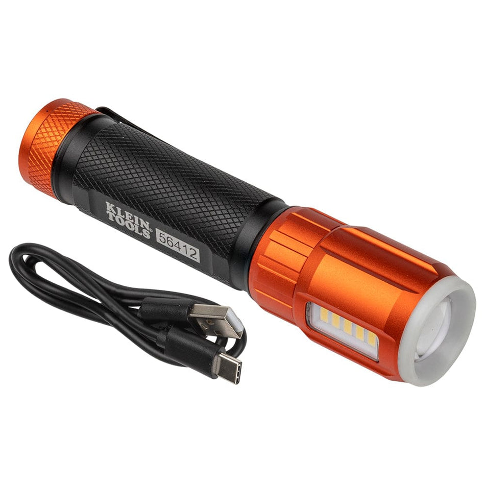 The image shows a cylindrical flashlight with a black textured body and orange ends, lying diagonally on a white surface. A black charging cable is coiled beside it. The flashlight has the words "KLEIN TOOLS 56412" printed on its side.