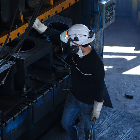 A person in a white hard hat, face mask, and protective gloves stands beside a large industrial machine in a factory setting, holding metal rods.