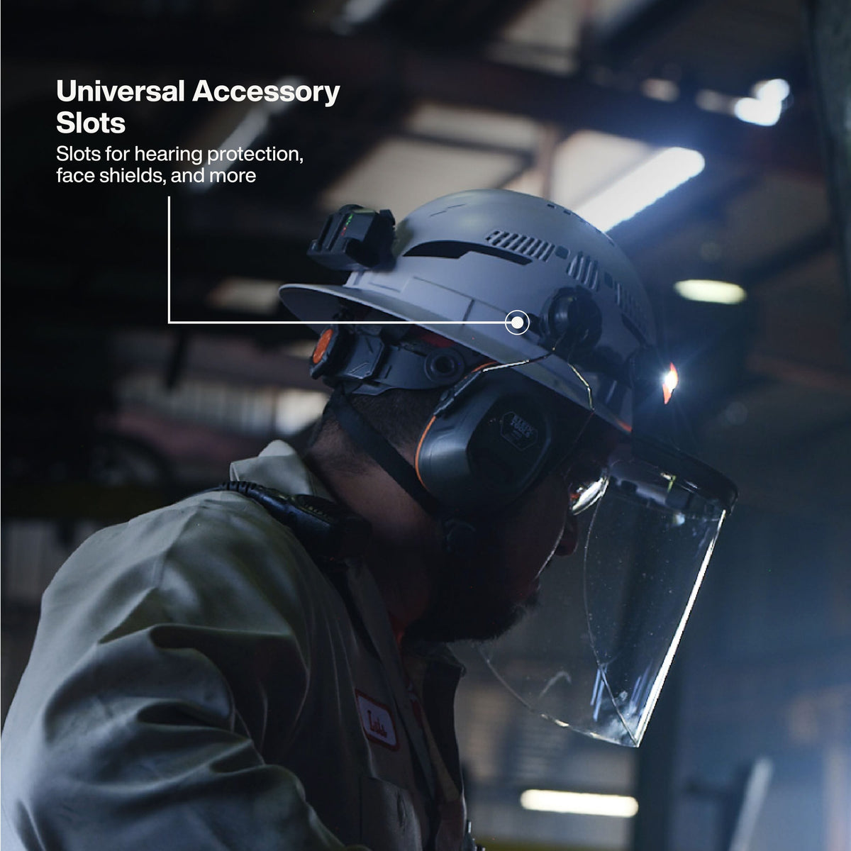 A person in a work environment wearing a gray hard hat with a clear face shield and ear protection. White text on the left reads 'Universal Accessory Slots' with a line pointing to the hard hat, indicating accessory slots.