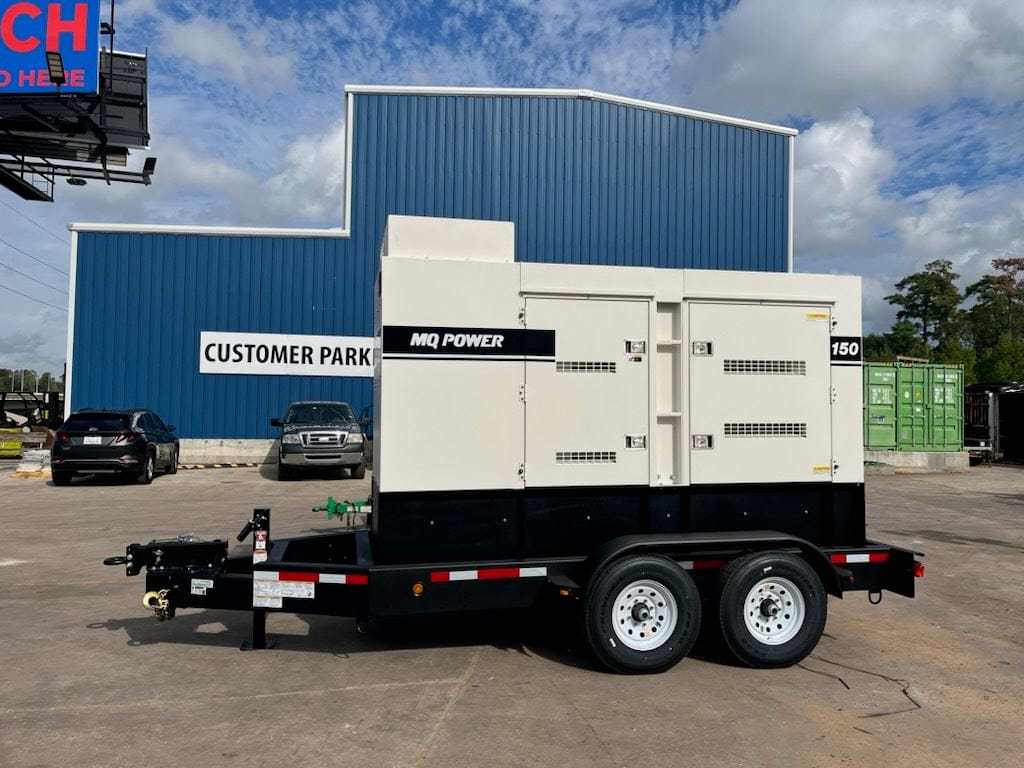  Large, light-colored generator with the "MQ Power" logo sits on a black trailer with two wheels. To the left, a sign reads "CUSTOMER PARKI" and beyond that are parked vehicles in front of a blue building.