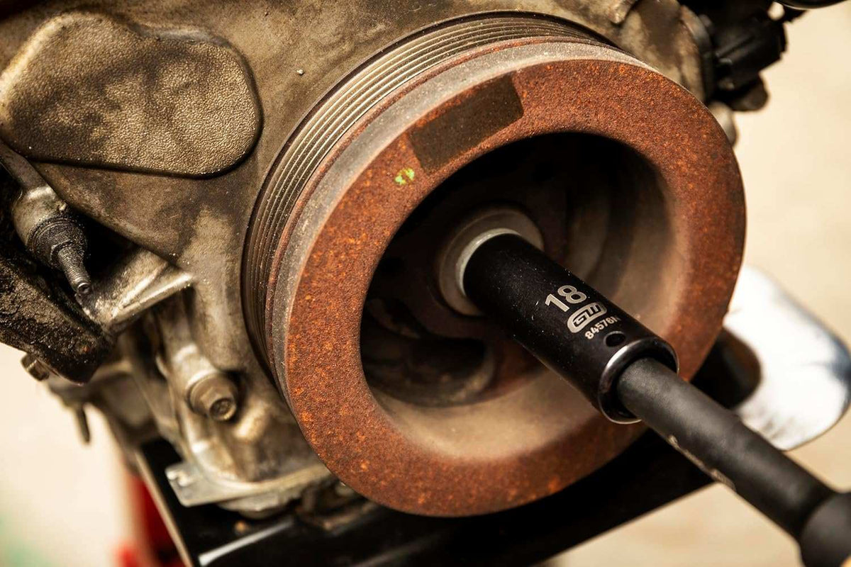 A GEARWRENCH 18mm deep impact socket (84576N) is shown attached to an extension bar, inserted into the central bolt of a rusty engine crankshaft pulley.