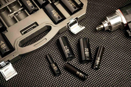 Several metric impact sockets (8mm, 15mm, 27mm, 28mm) from the GEARWRENCH 84935N set are laid out on a black mat next to the open blow-molded case and an impact wrench.