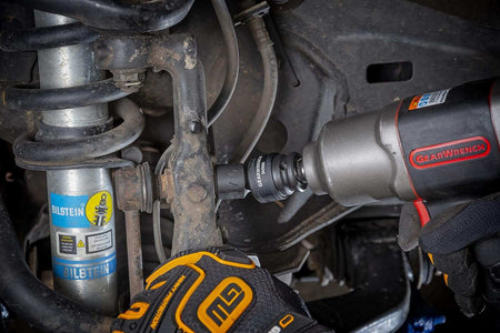 A gloved hand uses a GEARWRENCH 11/16-inch universal impact socket (84599N) with an impact wrench to remove a bolt from a vehicle's suspension assembly.