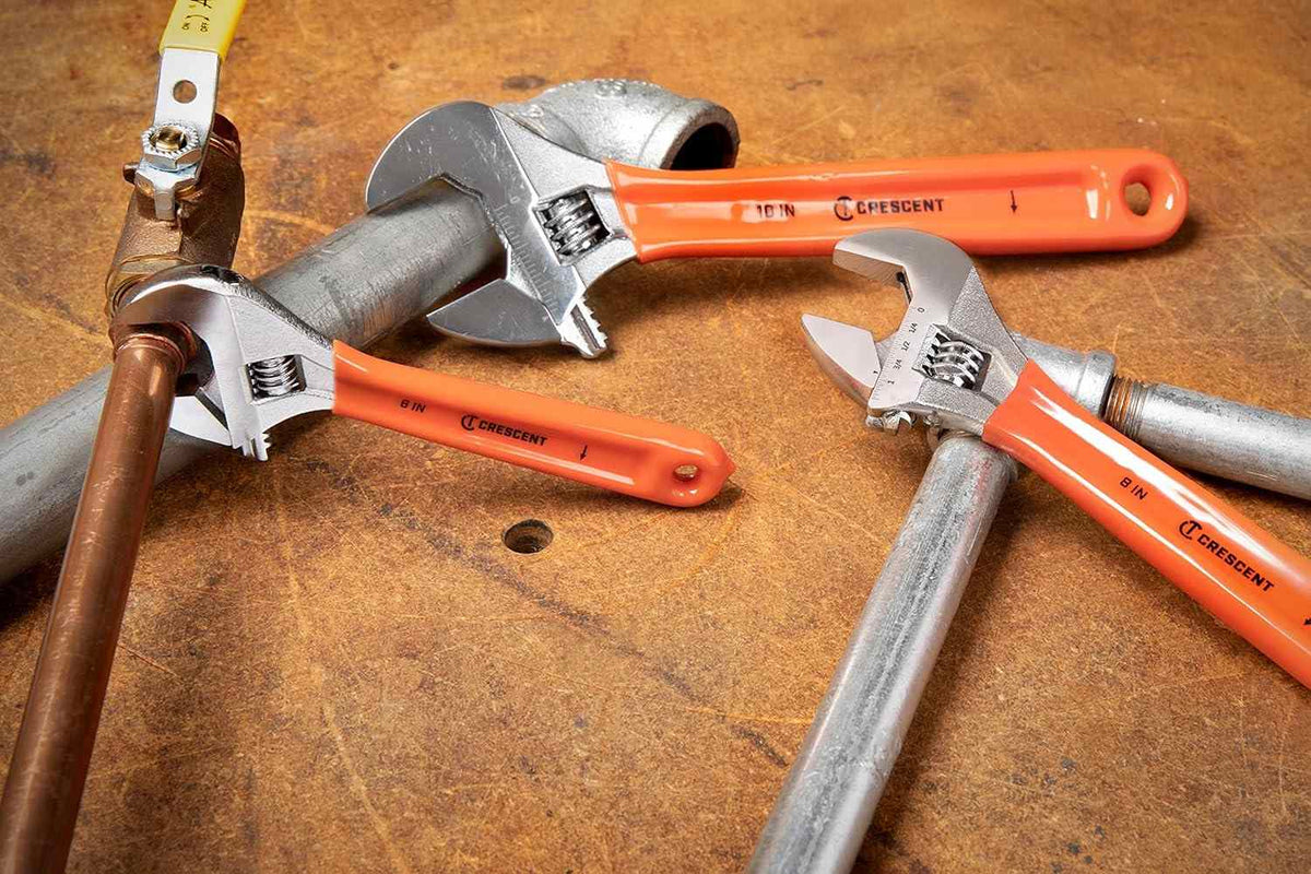 Three Crescent adjustable wrenches with orange cushion grips clamped onto different metal pipes (galvanized steel and copper) on a wooden workbench, demonstrating their use in plumbing.