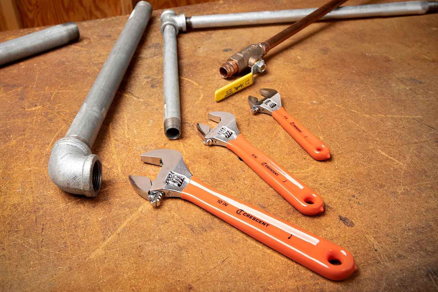 The Crescent 3-piece adjustable wrench set (10-inch, 8-inch, and 6-inch) with orange cushion grips laid out on a wooden surface next to various galvanized steel and copper plumbing pipes and a valve.