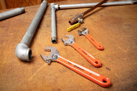 The Crescent 3-piece adjustable wrench set (10-inch, 8-inch, and 6-inch) with orange cushion grips laid out on a wooden surface next to various galvanized steel and copper plumbing pipes and a valve.