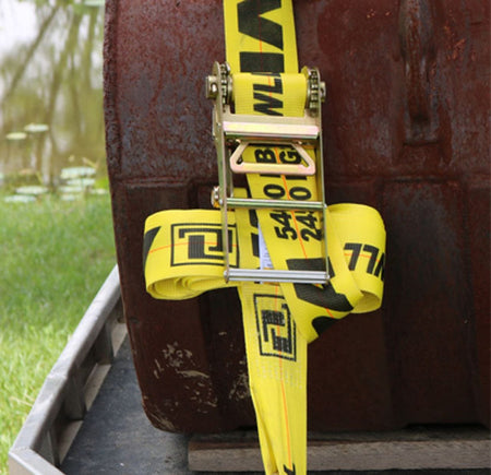 Close-up of the heavy-duty metal ratchet mechanism tightening the yellow 4-inch strap around a large, rusty metal tank on a trailer, demonstrating the strap's securement use.