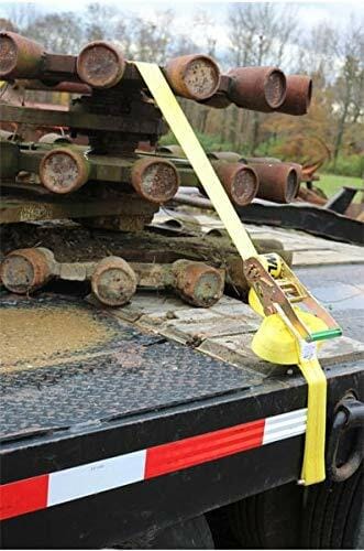 Action shot of the yellow ratchet strap securing metal pipes on a flatbed trailer, demonstrating the hardware's position over the cargo edge for industrial tie-down.