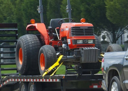 Full view of a yellow industrial ratchet strap used to securely anchor an orange utility tractor onto a flatbed trailer, demonstrating its application for heavy equipment.