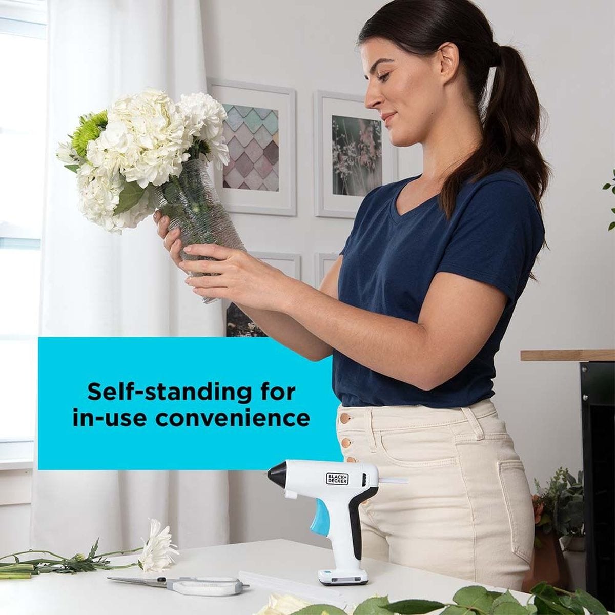 Woman examining a floral arrangement with the BLACK+DECKER 4V MAX cordless hot glue gun securely resting on its self-standing base on the table beside her.
