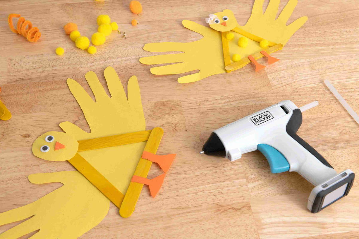 The BLACK+DECKER BCGL115FF cordless craft glue gun laying on a wooden table next to completed children's crafts made from construction paper and popsicle sticks.