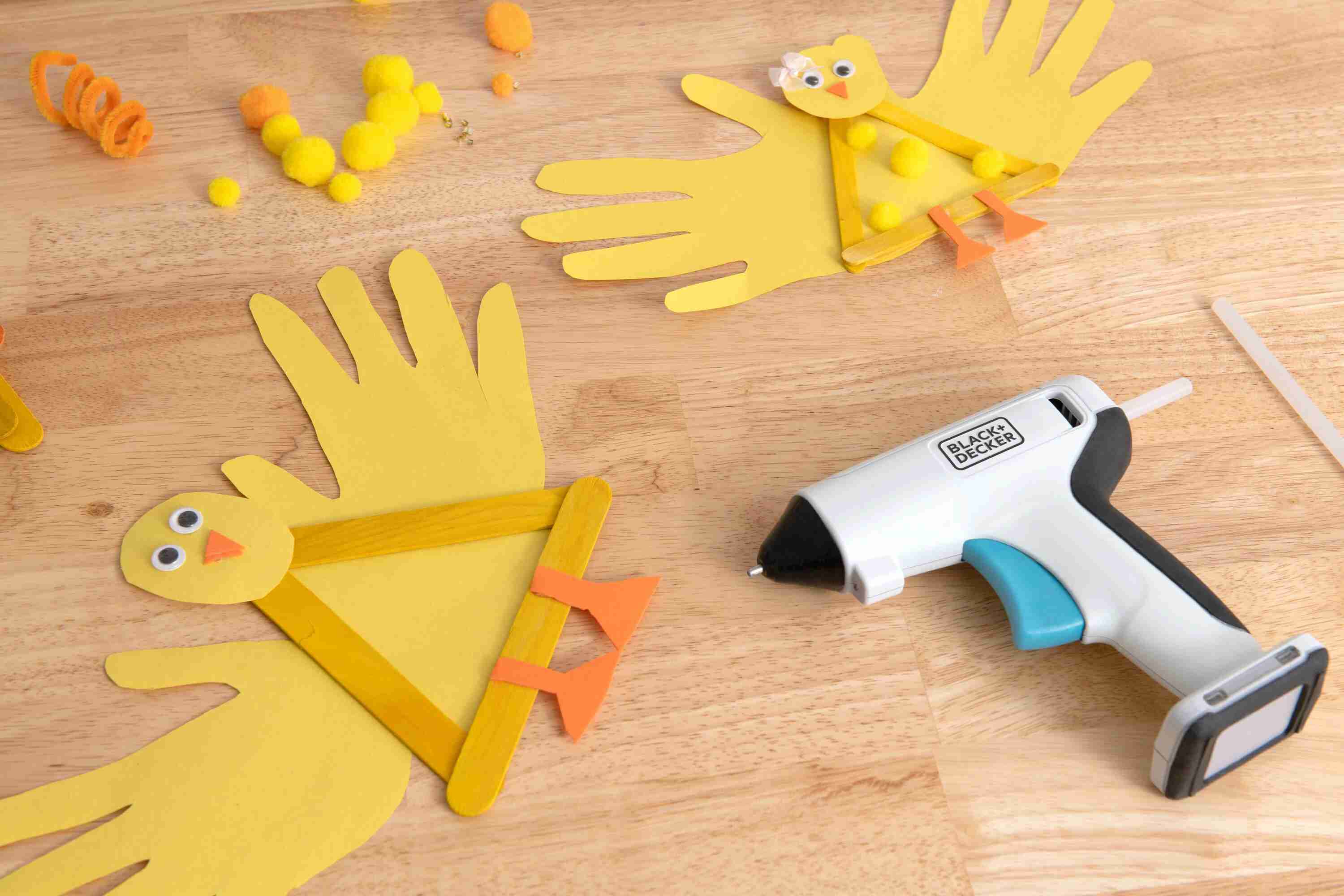 The BLACK+DECKER BCGL115FF cordless craft glue gun laying on a wooden table next to completed children's crafts made from construction paper and popsicle sticks.