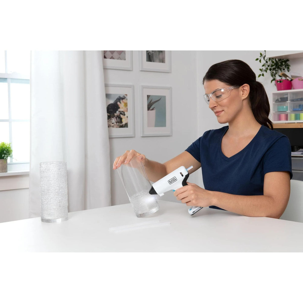 Woman wearing safety glasses using the portable BLACK+DECKER cordless hot glue gun to apply adhesive to a glass vase for a home DIY project.