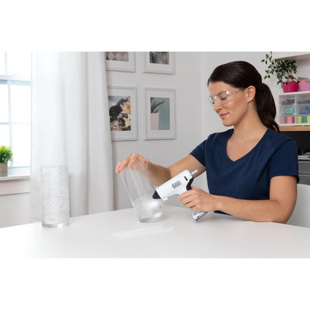 Woman wearing safety glasses using the portable BLACK+DECKER cordless hot glue gun to apply adhesive to a glass vase for a home DIY project.