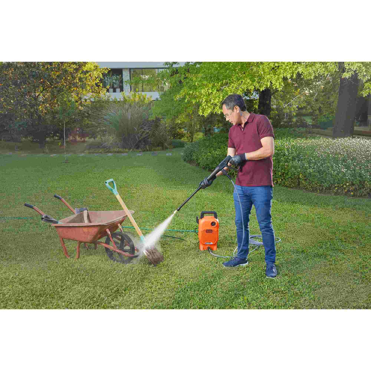 A person uses a BLACK+DECKER pressure washer to clean a red wheelbarrow in a grassy backyard. The orange pressure washer is placed nearby, connected to a hose and power source.
