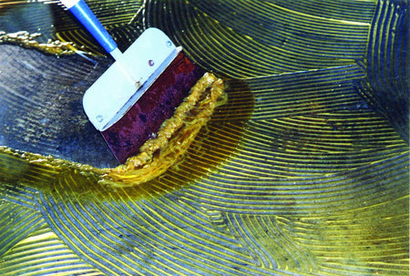 Close-up action shot showing a scraper lifting a strip of thick, dissolved yellow floor mastic with trowel ridges from a concrete subfloor.