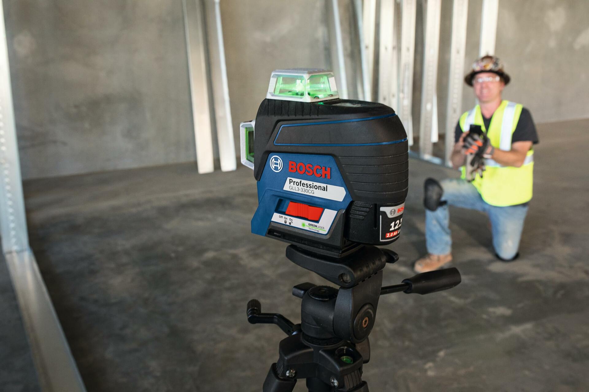 Bosch laser level on tripod projecting green laser lines, with construction worker kneeling nearby in an unfinished interior using alignment tools