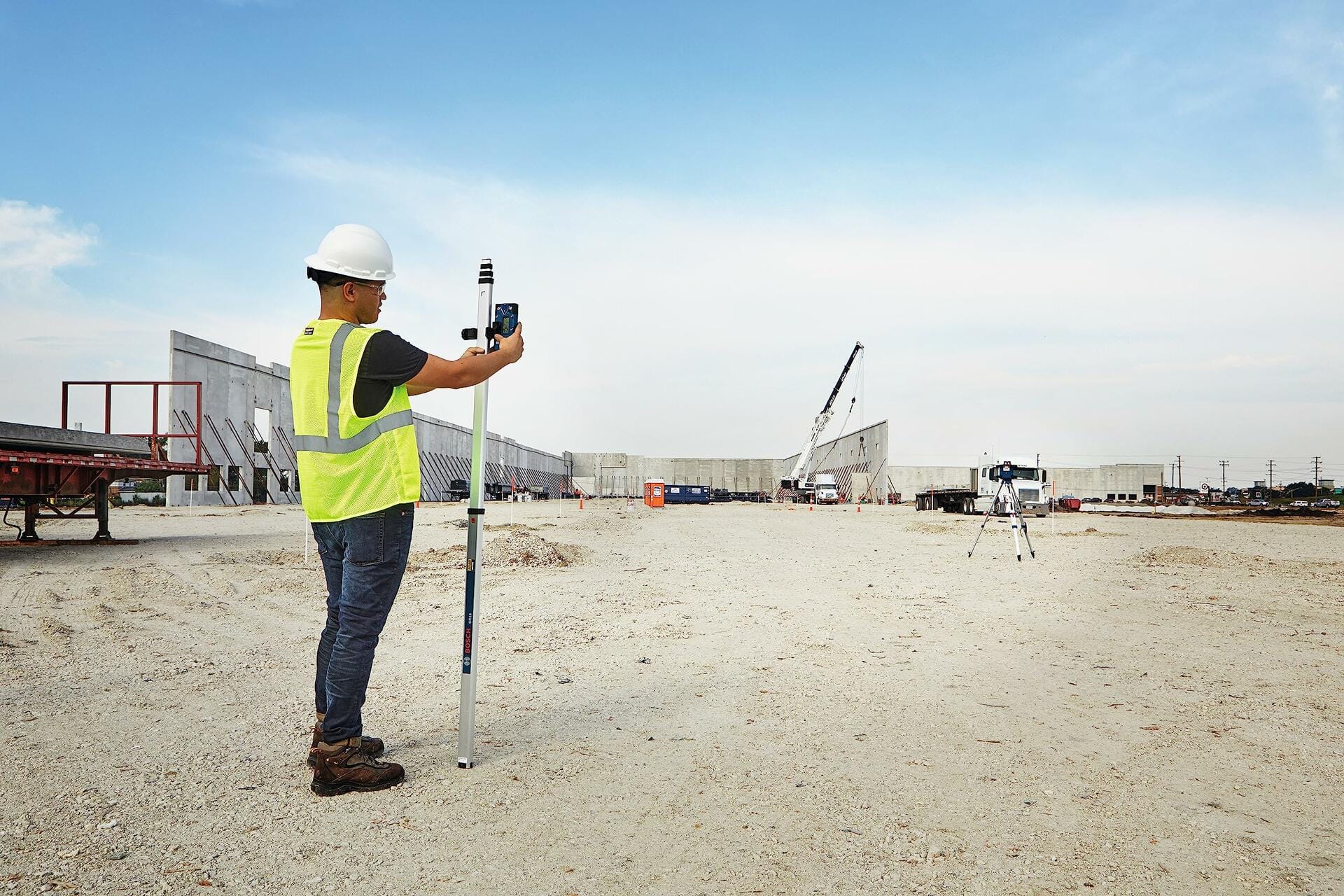Construction worker in a hard hat and safety vest operating the BOSCH LR40 Receiver mounted on a grade rod to check elevation across a large, open construction site with a rotary laser visible on a tripod in the distance.