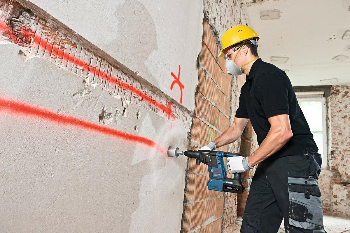Worker using power drill on plaster wall with red spray-painted markings. Wearing hard hat, dust mask, safety glasses, and reinforced work pants. Exposed brick and debris visible in renovation site.