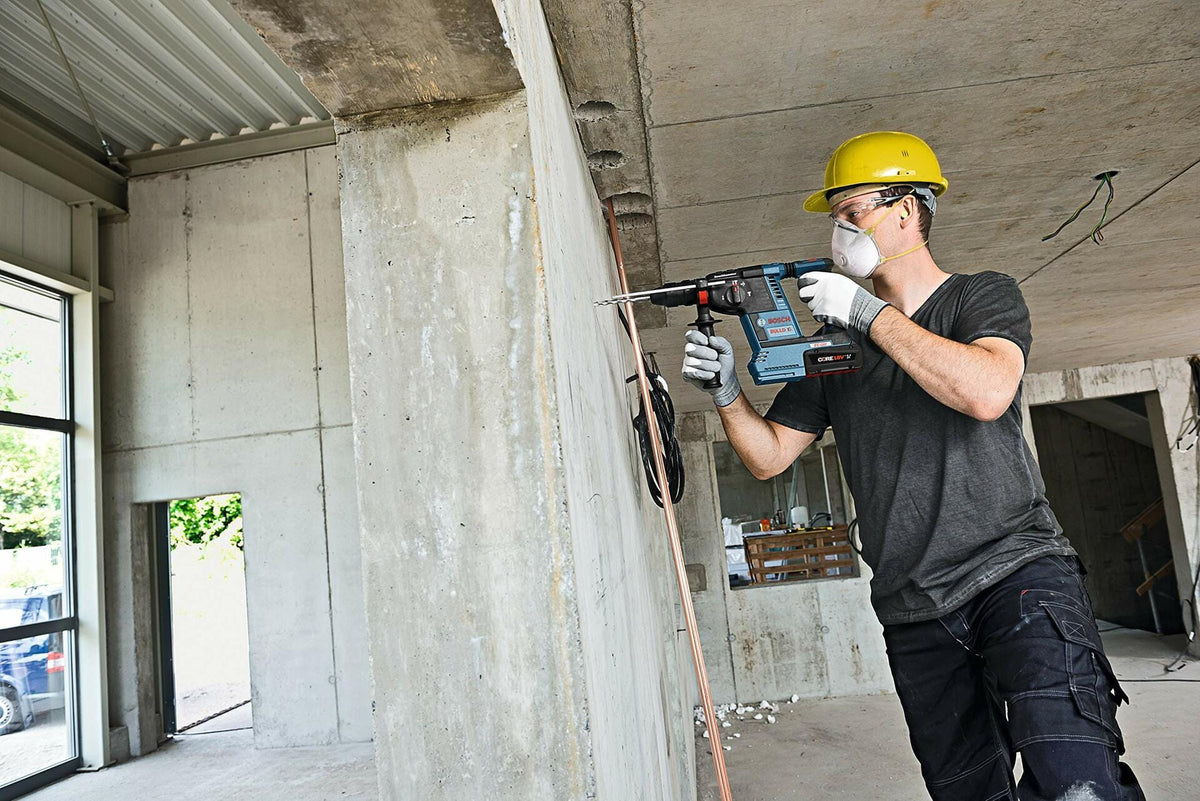 Worker using rotary hammer drill on concrete wall in unfinished building. Wearing hard hat, dust mask, safety glasses, and gloves. Electrical wiring visible overhead.