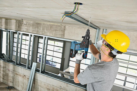 Worker using power drill on ceiling with exposed electrical wires. Wearing hard hat, safety glasses, ear protection, and gloves. Standing on platform in unfinished building with concrete walls.